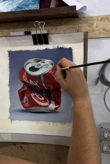 Una mano pinta una lata de Coca-Cola arrugada sobre una mesa de trabajo.